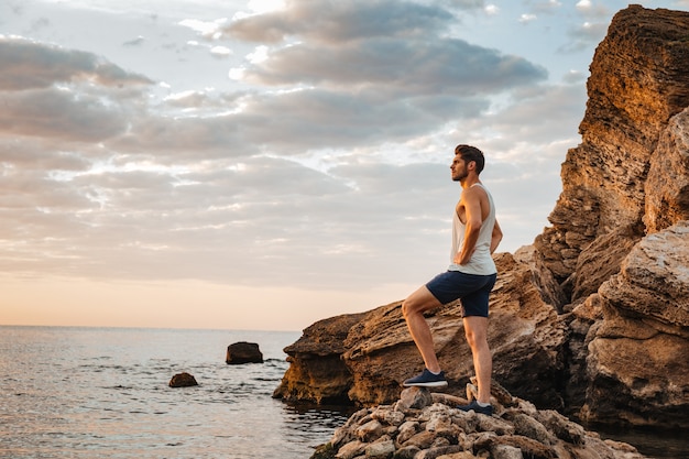 Giovane atleta bello in piedi sulla spiaggia rocciosa in riva al mare