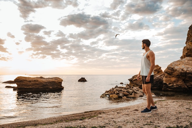 Giovane atleta bello in piedi sulla spiaggia rocciosa in riva al mare