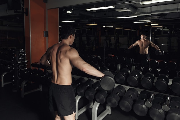 Молодой красивый спортсмен тренирует его плечи с тренажерным залом гантелей у зеркала.
