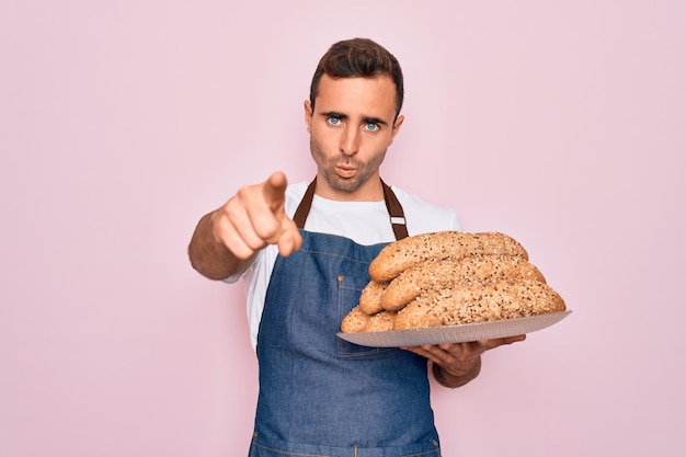 Young handsome baker man with blue eyes wearing apron holding tray with wholmeal bread pointing with finger to the camera and to you hand sign positive and confident gesture from the front