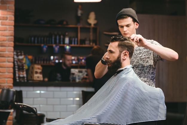 Young handsome barber making haircut of attractive bearded man in barbershop