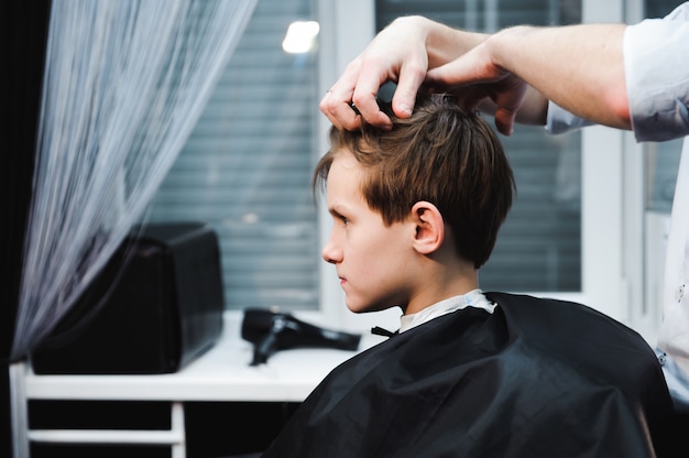 Young handsome barber making haircut of cute boy in barbershop