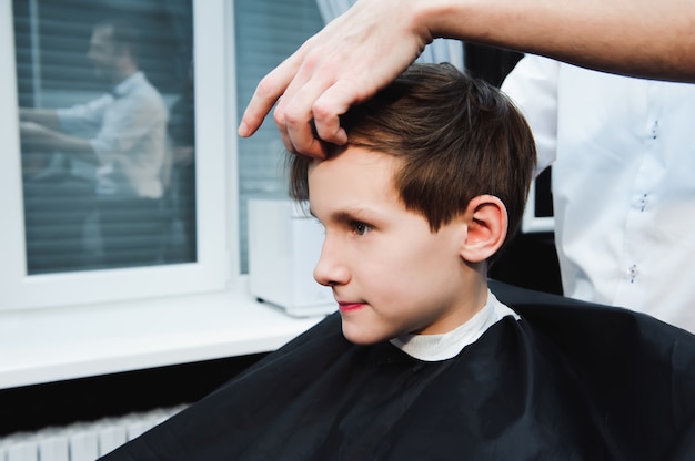Young handsome barber making haircut of cute boy in barbershop