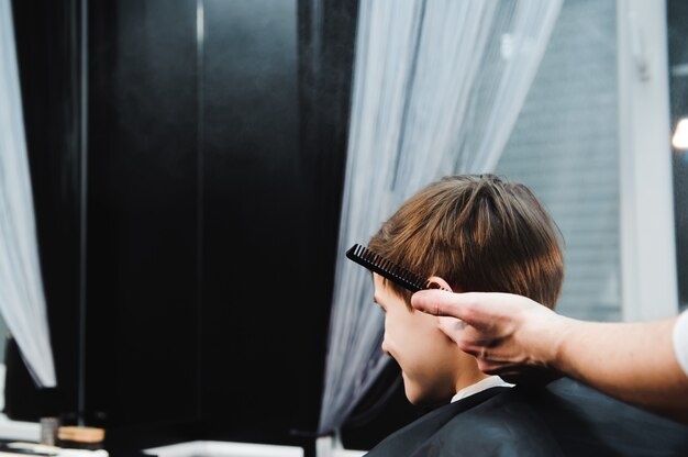 Young handsome barber making haircut of cute boy in barbershop