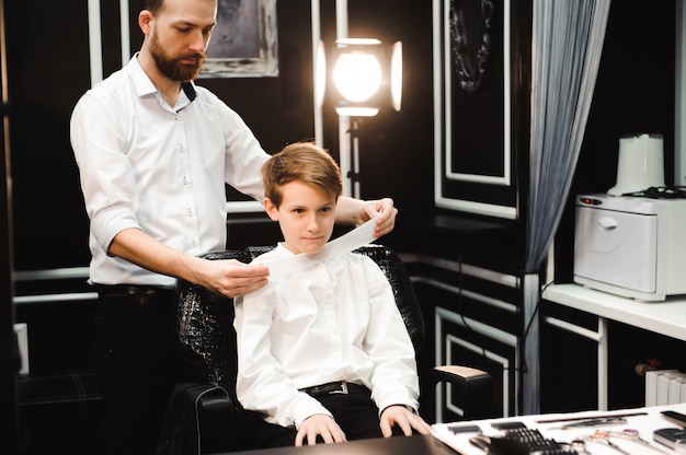 Young handsome barber making haircut of cute boy in barbershop.