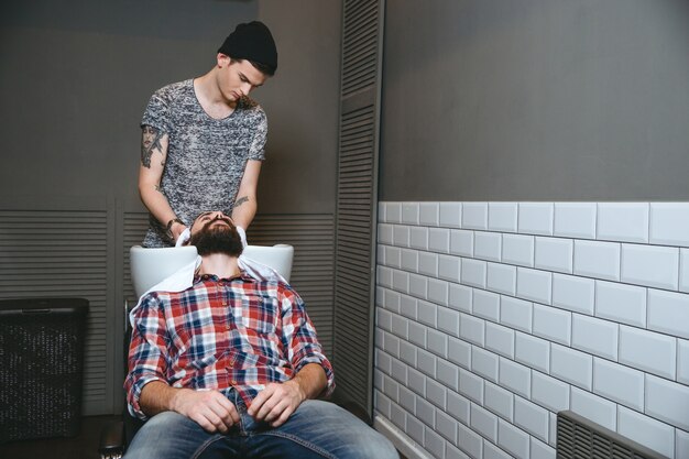 Young handsome barber with tatoo in black hat washing hair of client with beard in plaid shirt and jeans in barbershop