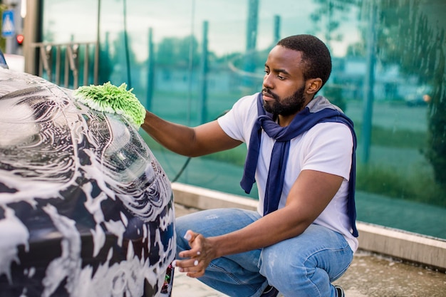 Young handsome bearded african man in tshirt and jeans cleaning\
his luxury electric gray car outdoors at self car wash station\
using green rag and soap foam