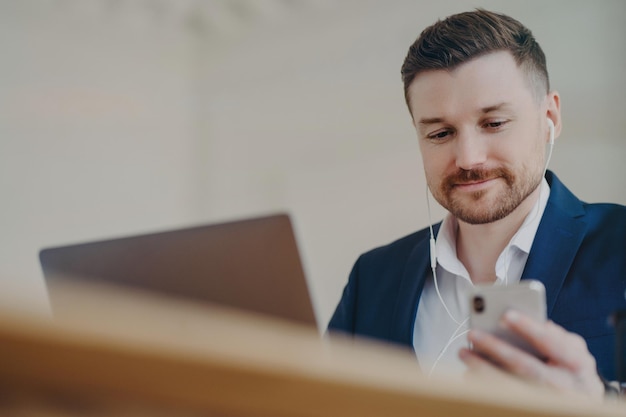 Young handsome bearded businessman wearing earphones having online meeting while sitting in front of laptop male manager in stylish suit using cell phone while sitting behind office desk