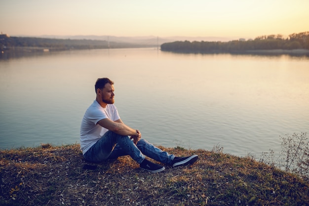 Foto giovane uomo biondo caucasico barbuto bello che si siede sulla scogliera e che esamina tramonto