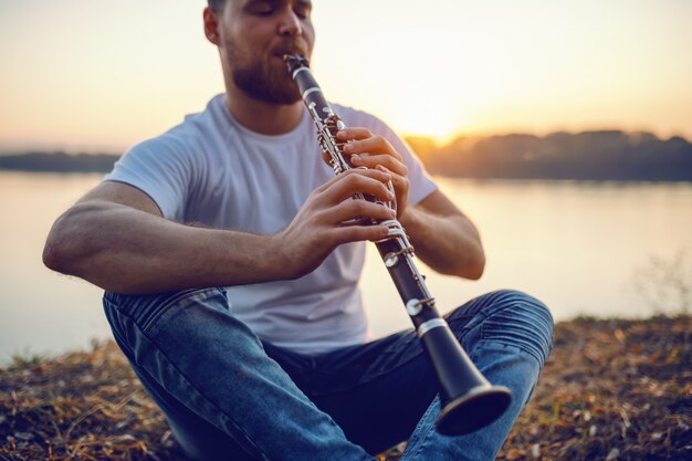 Young handsome bearded caucasian blond man sitting on cliff and playing clarinet.