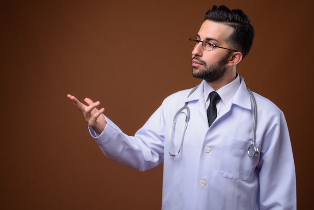 Young handsome bearded Iranian man doctor against brown wall