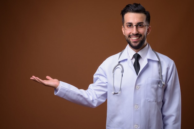 Young handsome bearded Iranian man doctor against brown wall