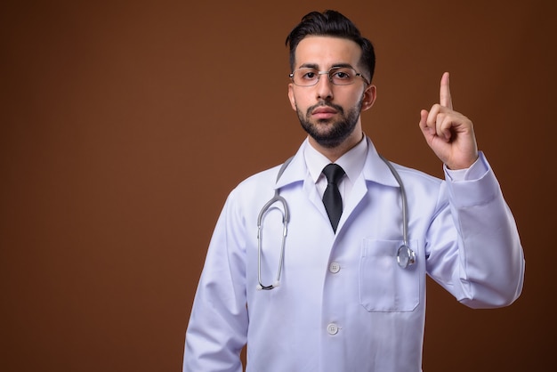 Young handsome bearded Iranian man doctor against brown wall