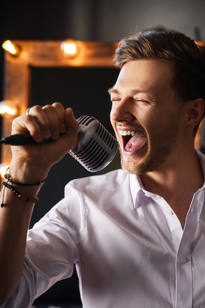 Young handsome bearded man is singing loudly into a microphone with his eyes closed at home with a makeup mirror behind his back