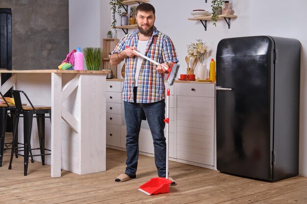 Young handsome bearded man in the kitchen wearing checkered shirt tries to deal with a dustpan and a...