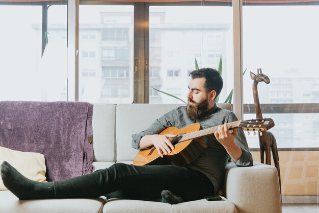 Young handsome bearded Man playing acoustic spanish guitar sitting on the sofa in the living room. Learning to play a new instrument. New habit concept, skills and hobby. Modern home trendy clothes