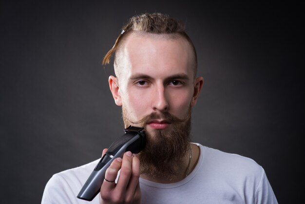 Young handsome bearded man shaves thick beard with trimmer in studio