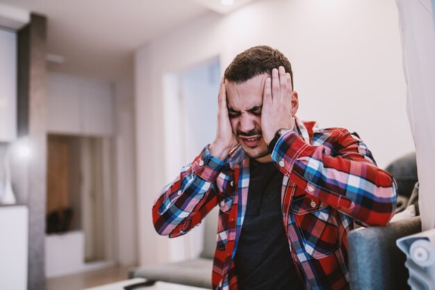 Young handsome bearded man sitting on sofa in living room, holding head and having headache.