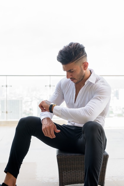 Young handsome bearded Persian businessman checking the time against view the city