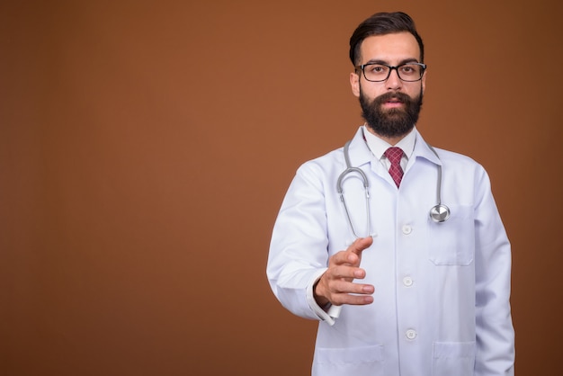 Young handsome bearded Persian man doctor on brown wall