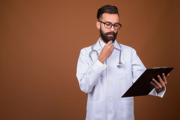 Young handsome bearded Persian man doctor on brown wall