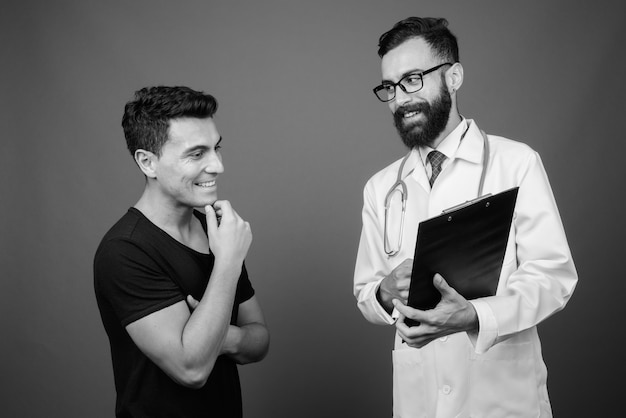 young handsome bearded Persian man doctor with young Hispanic man patient against gray wall in black and white