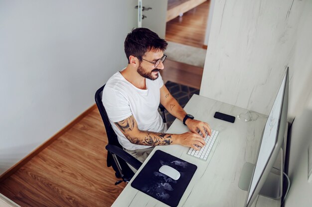 Young handsome bearded smiling freelancer sitting in his home office and typing important things about new project.