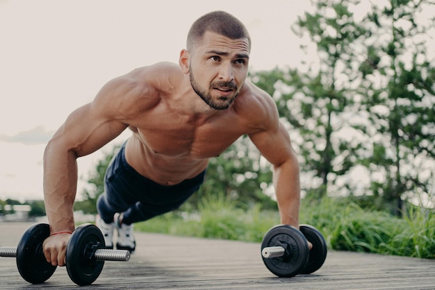 Young handsome bearded sportsman works out outdoor poses in plank with barbells has strong body muscular arms Bodybuilder does exercises in open air performs weight lifting push up exercises