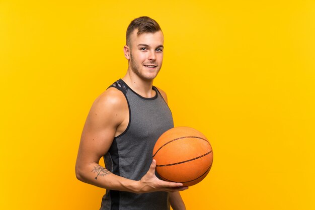 Young handsome blonde man holding a basket ball over yellow wall smiling a lot