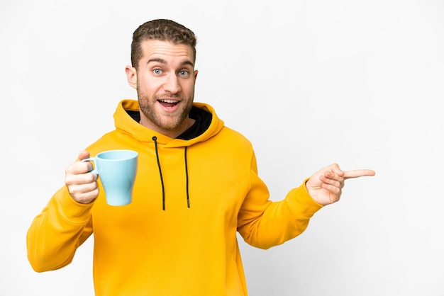 Young handsome blonde man holding cup of coffee over isolated background surprised and pointing finger to the side