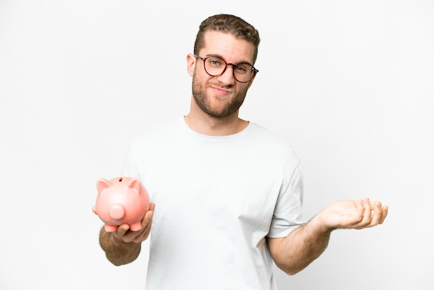 肩を持ち上げながら疑問のジェスチャーを作る piggybank を保持している若いハンサムな金髪の男