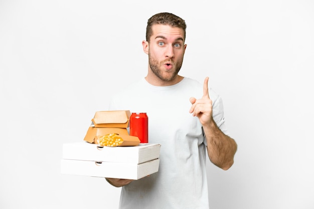 Young handsome blonde man holding pizzas and burgers over isolated background intending to realizes the solution while lifting a finger up