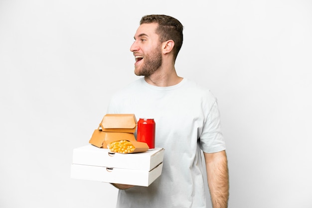 Young handsome blonde man holding pizzas and burgers over isolated background laughing in lateral position