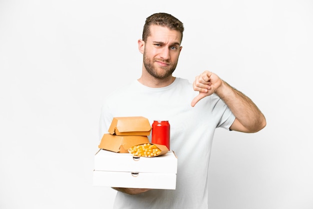 Young handsome blonde man holding pizzas and burgers over isolated background showing thumb down with negative expression