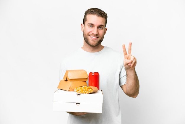 Young handsome blonde man holding pizzas and burgers over isolated background smiling and showing victory sign