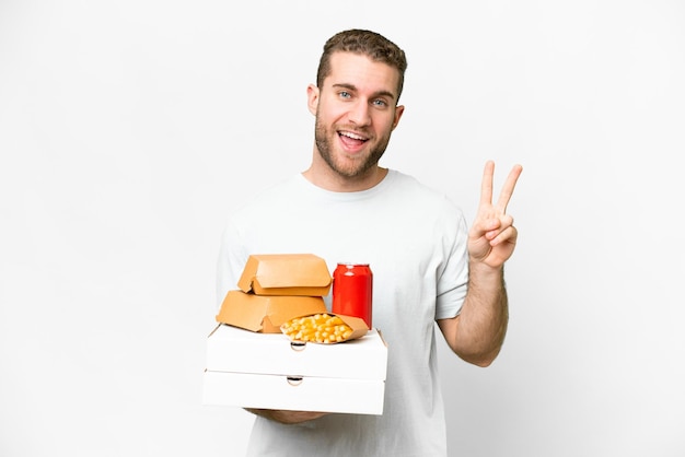 Young handsome blonde man holding pizzas and burgers over isolated background smiling and showing victory sign