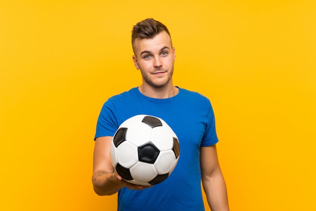 Young handsome blonde man holding a soccer ball over isolated yellow background with happy expression