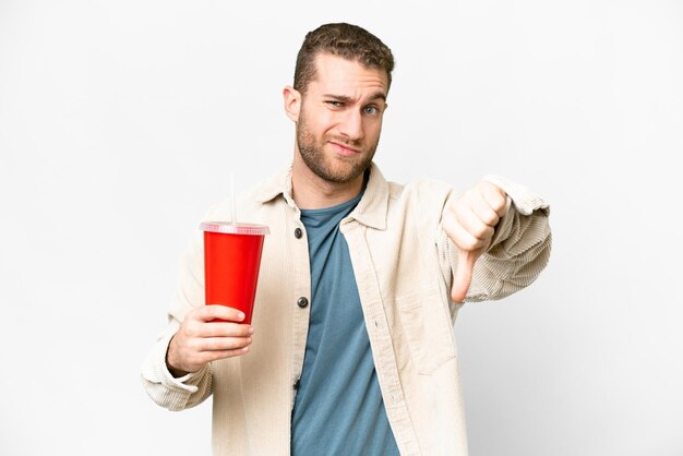 Young handsome blonde man holding soda over isolated white background showing thumb down with negative expression