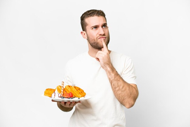 Photo young handsome blonde man holding waffles over isolated white background having doubts while looking up