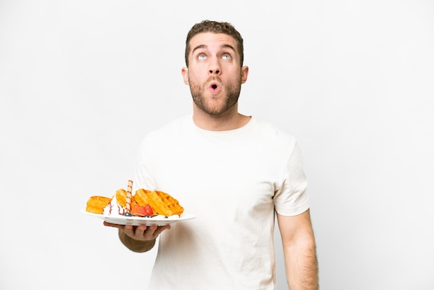 Young handsome blonde man holding waffles over isolated white background looking up and with surprised expression