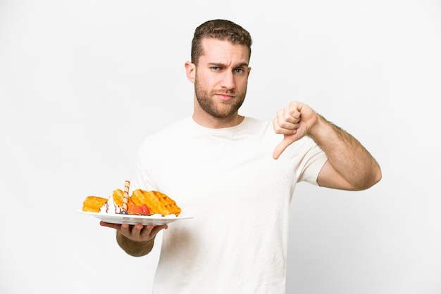 Young handsome blonde man holding waffles over isolated white background showing thumb down with negative expression