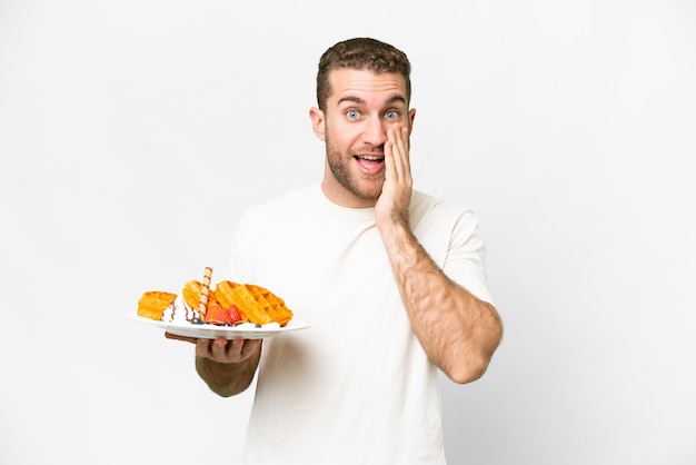 Young handsome blonde man holding waffles over isolated white background with surprise and shocked facial expression