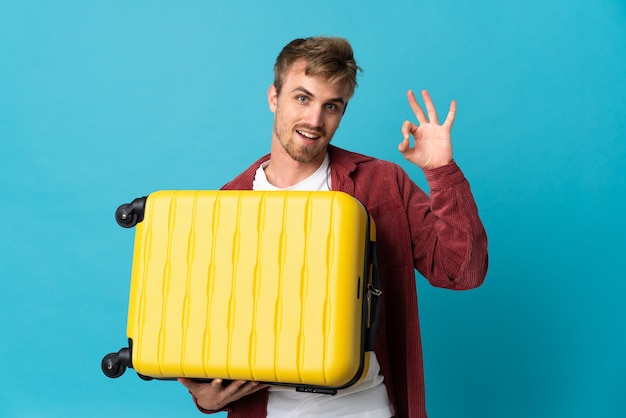Young handsome blonde man isolated on blue background in vacation with travel suitcase and making OK sign