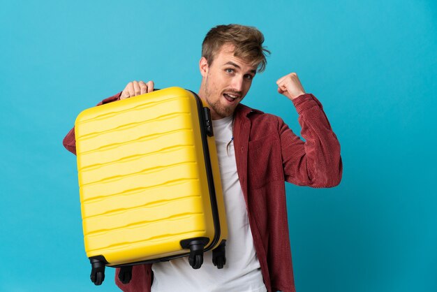 Young handsome blonde man isolated on blue background in vacation with travel suitcase