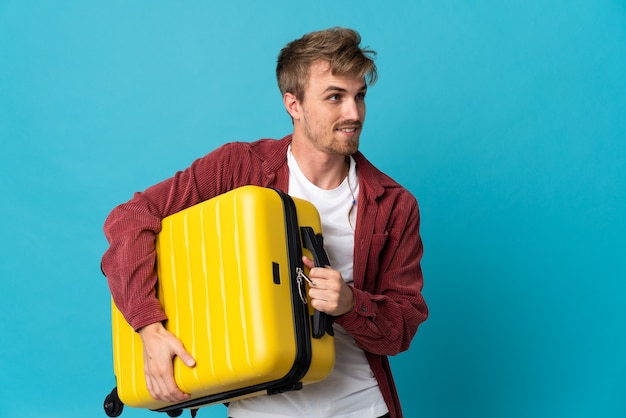 Young handsome blonde man isolated on blue background in vacation with travel suitcase