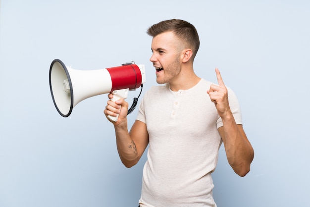 Young handsome blonde man over isolated blue wall shouting through a megaphone