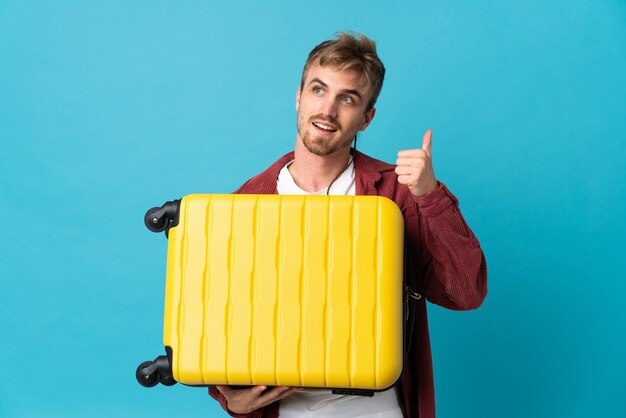 Young handsome blonde man isolated on blue wall in vacation with travel suitcase and with thumb up