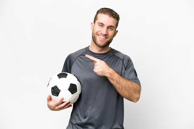 Young handsome blonde man over isolated white background with soccer ball and pointing to the lateral