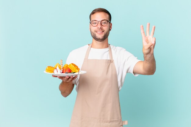 젊고 잘생긴 금발 남자가 웃고 친절하게 보이고 3번을 보여줍니다. 요리 와플 개념