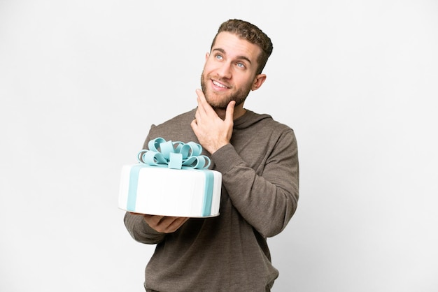 Young handsome blonde man with a big cake over isolated white background looking up while smiling